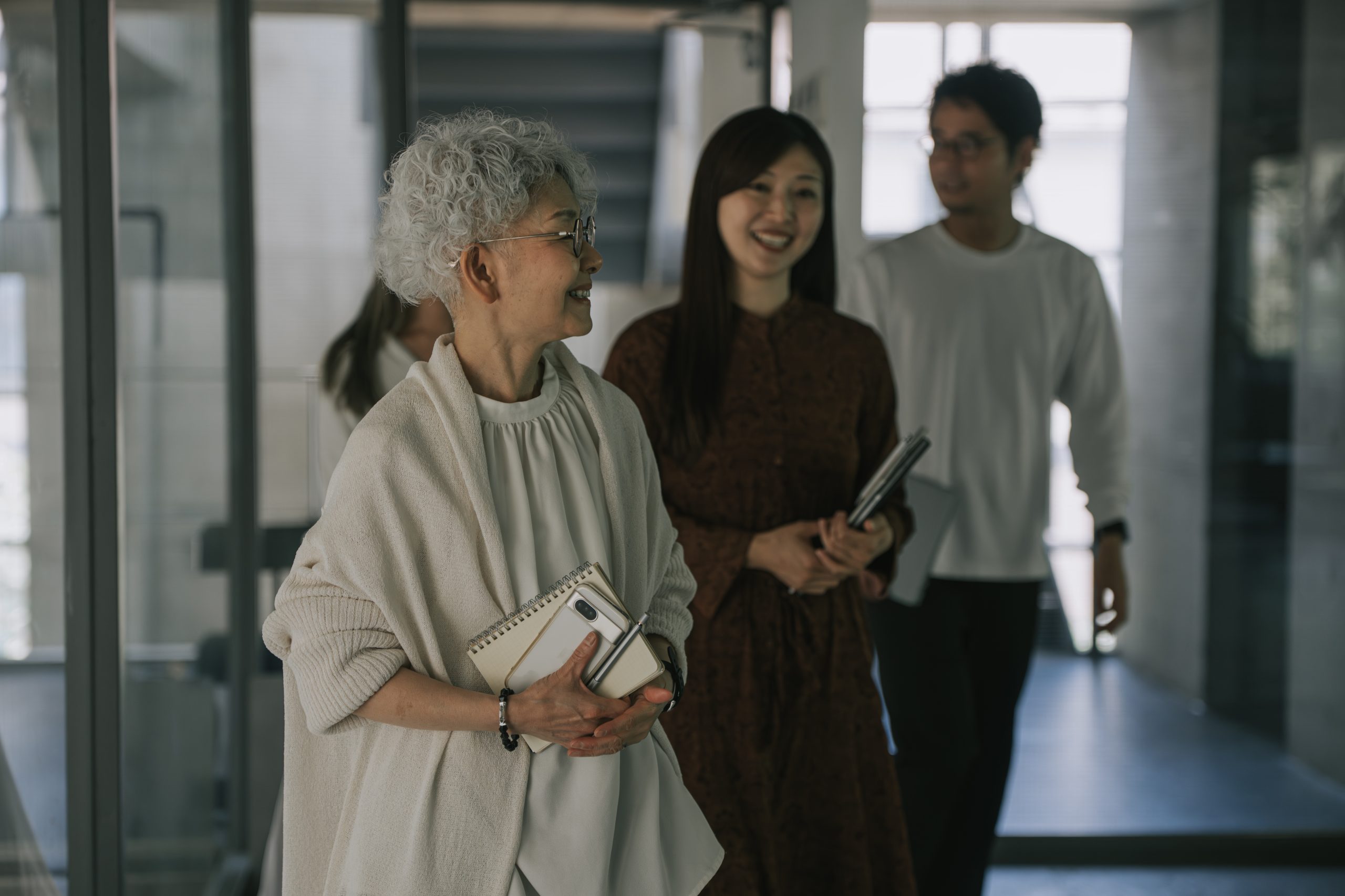 Smiling Senior Japanese Woman Walking with Younger Coworkers in Modern Office Hallway